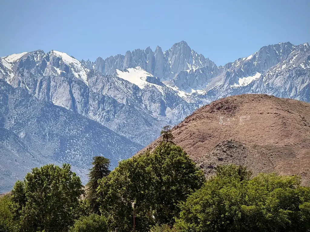 The jagged granite crest of Mt. Whitney above green trees and a brown foothill