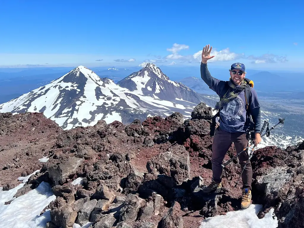 Luc waving on the rocky summit of South Sister, with snow-covered peaks behind him